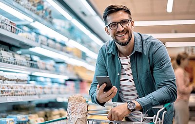 Junger Mann mit Bart und Brille stützt sich fröhlich auf Einkaufswagen in Supermarkt und hat sein Smartphone in der Hand