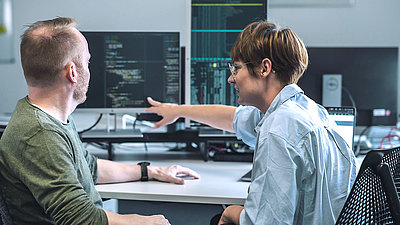 Ein IT-Spezialist und eine IT-Spezialistin beraten sich vor mehreren Monitoren mit Programmiersprache zu IT-Maßnahmen